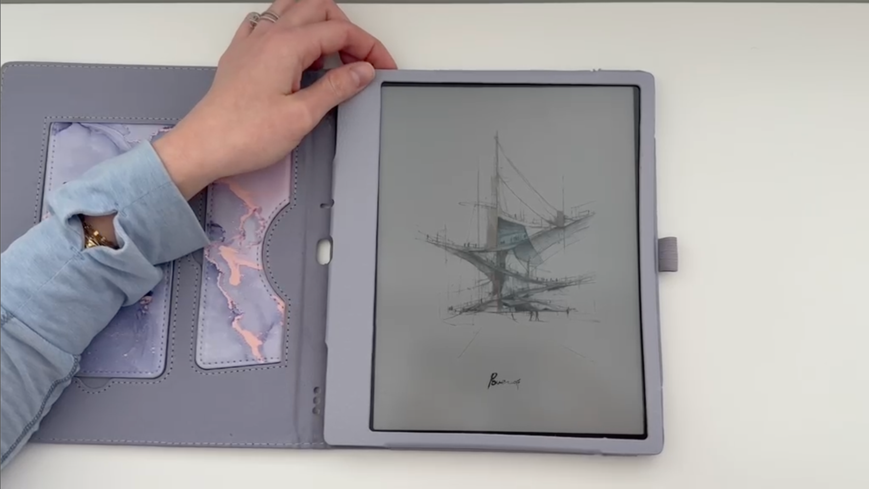 Hand opening a case to reveal an e-reader displaying an illustration on a white background. Finger pressing the on button at the top left.